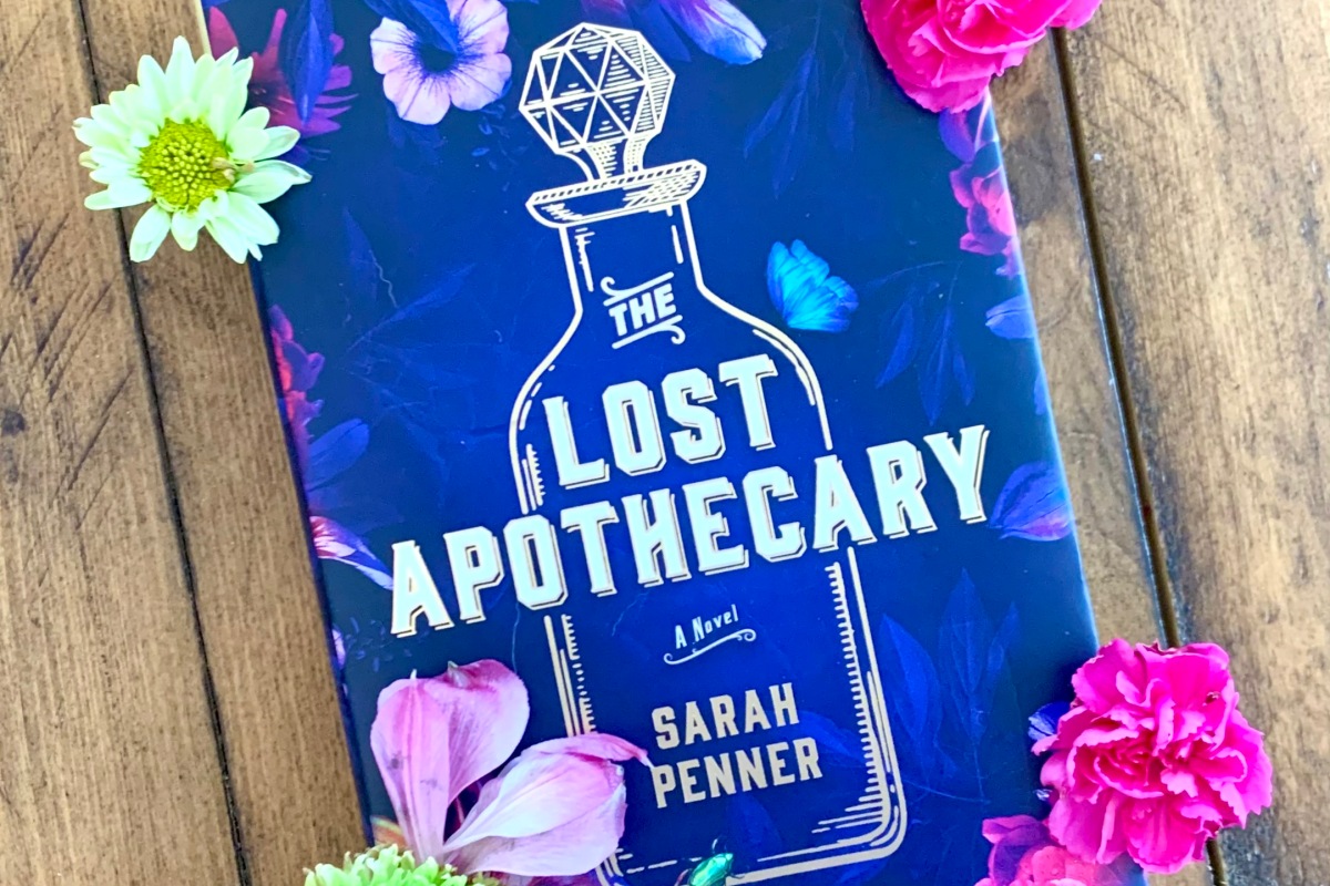 A copy of The Lost Apothecary by Sarah Penner, a purple hardback book with a bottle and flowers on the cover, with live flowers surrounding it that are pink, green, and white
